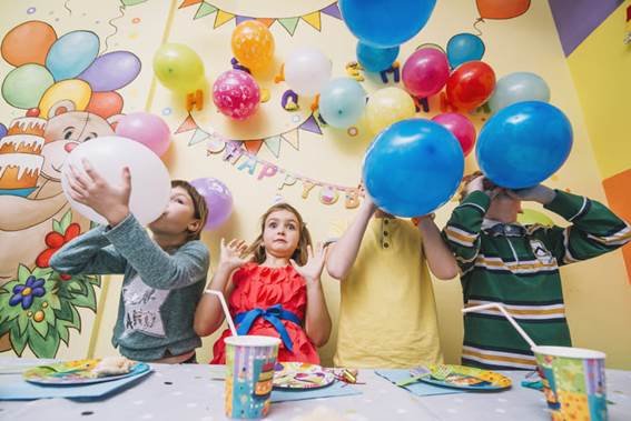 Nesta imagem, amiguinhos estao enchendo balões, para sua festinha de aniversario, com uma mesa bem colorida e bem divertida.